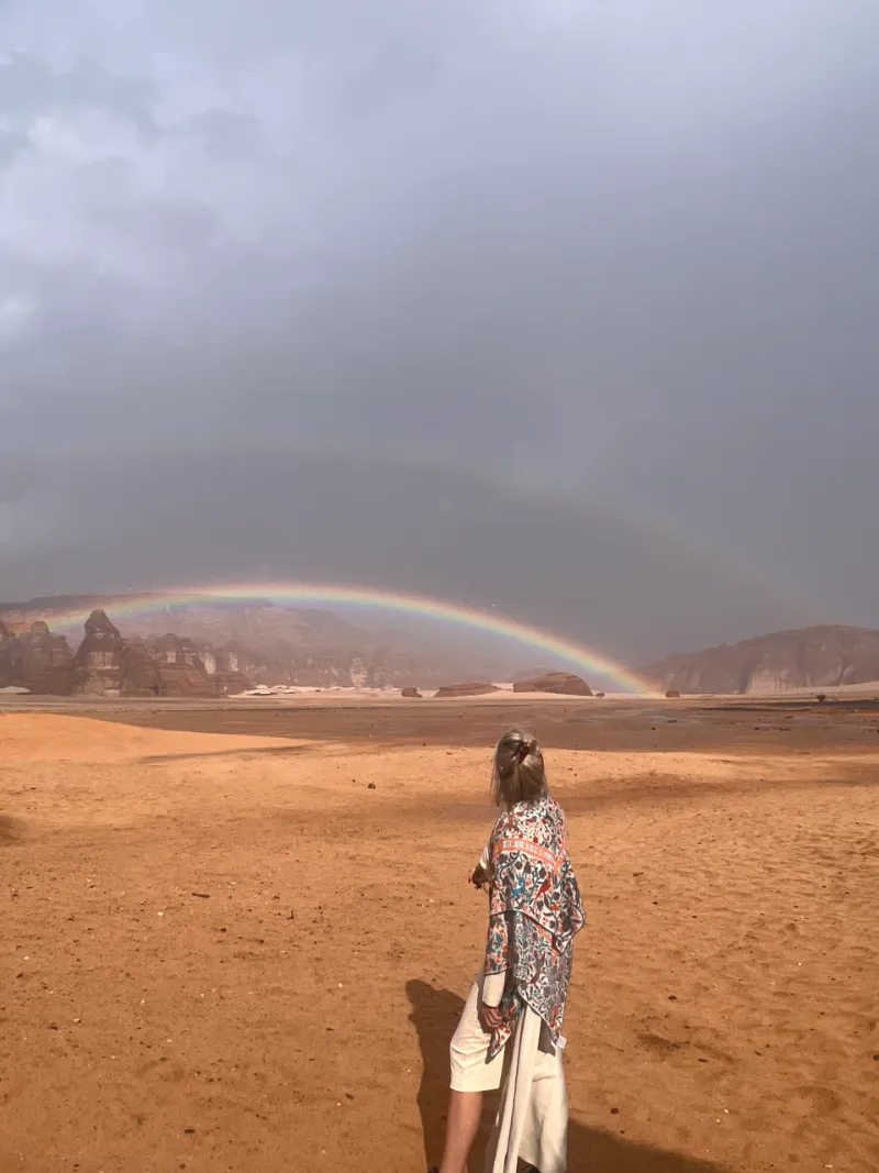 A figure standing in the desert under a rainbow