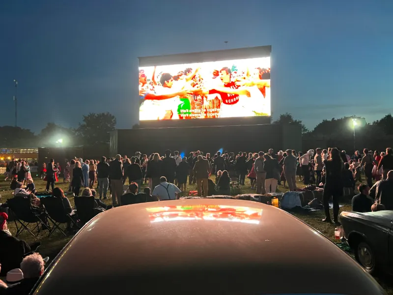 A giant outdoor screen at night with a standing crowd and a vintage car in front