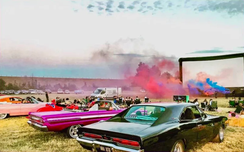 Vintage American cars with pink and blue smoke at an outdoor screen
